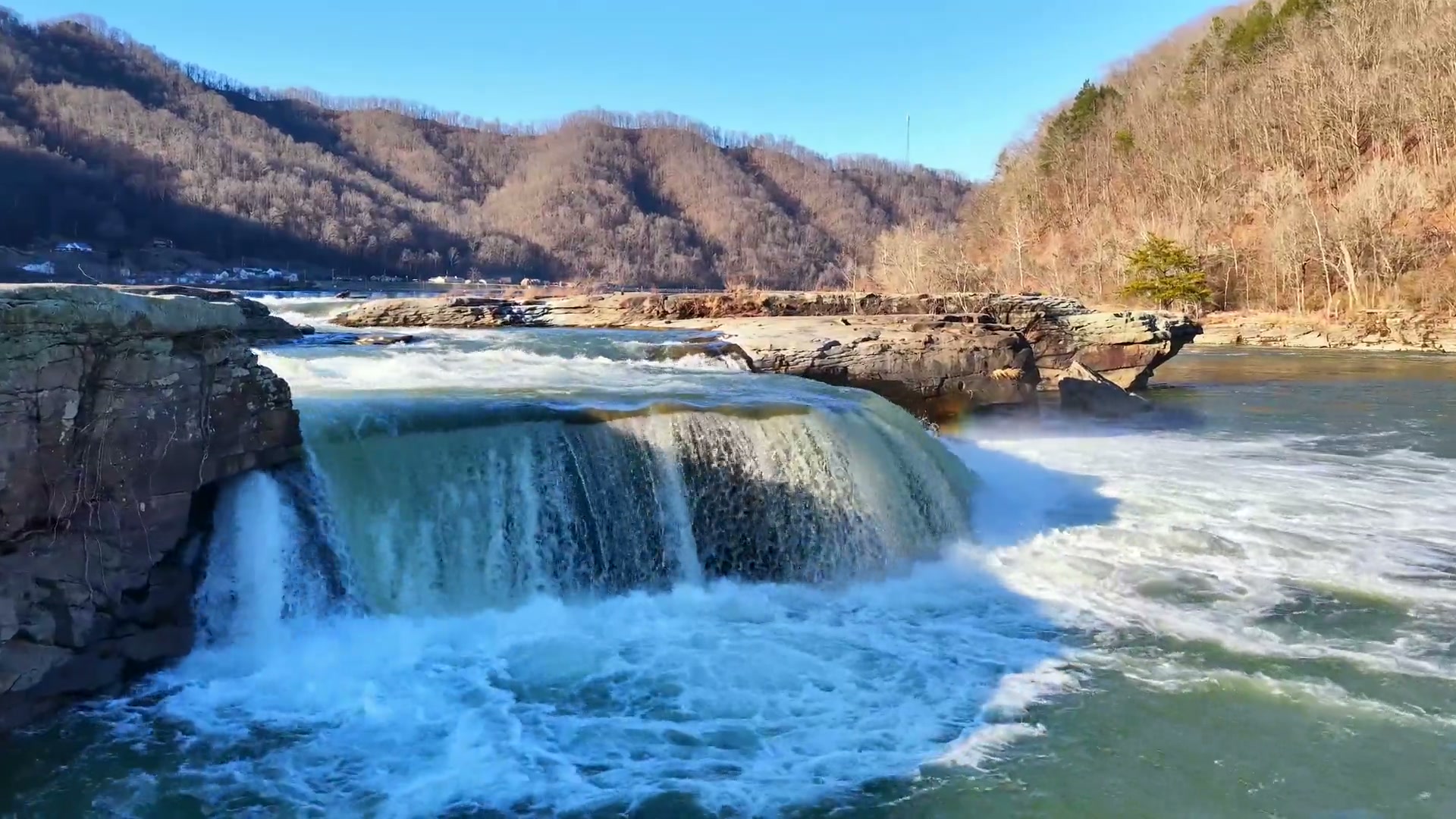 Aerial view of a powerful waterfall in a sunlit river gorge, filmed by Horizon Heights Productions.